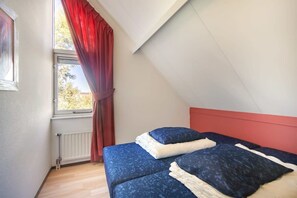 A bedroom with a sloped ceiling, a window with red curtains, and a bed with blue bedding.