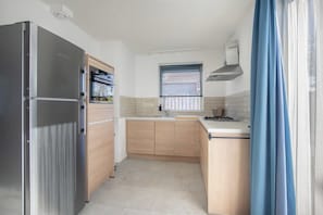 Een kleine keuken met een roestvrijstalen koelkast, houten kastjes, een raam met zonwering en een blauw gordijn.