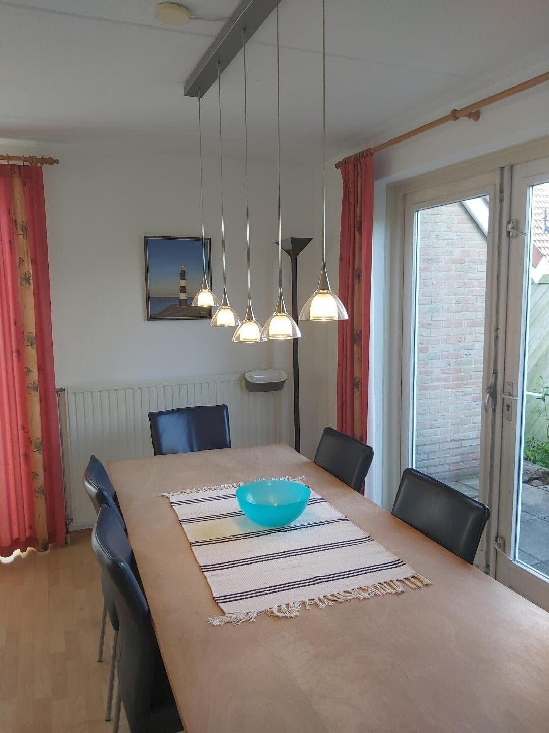 A dining area with a table, chairs, a blue bowl, hanging pendant lights, a painting, and a window with a view of a brick wall.