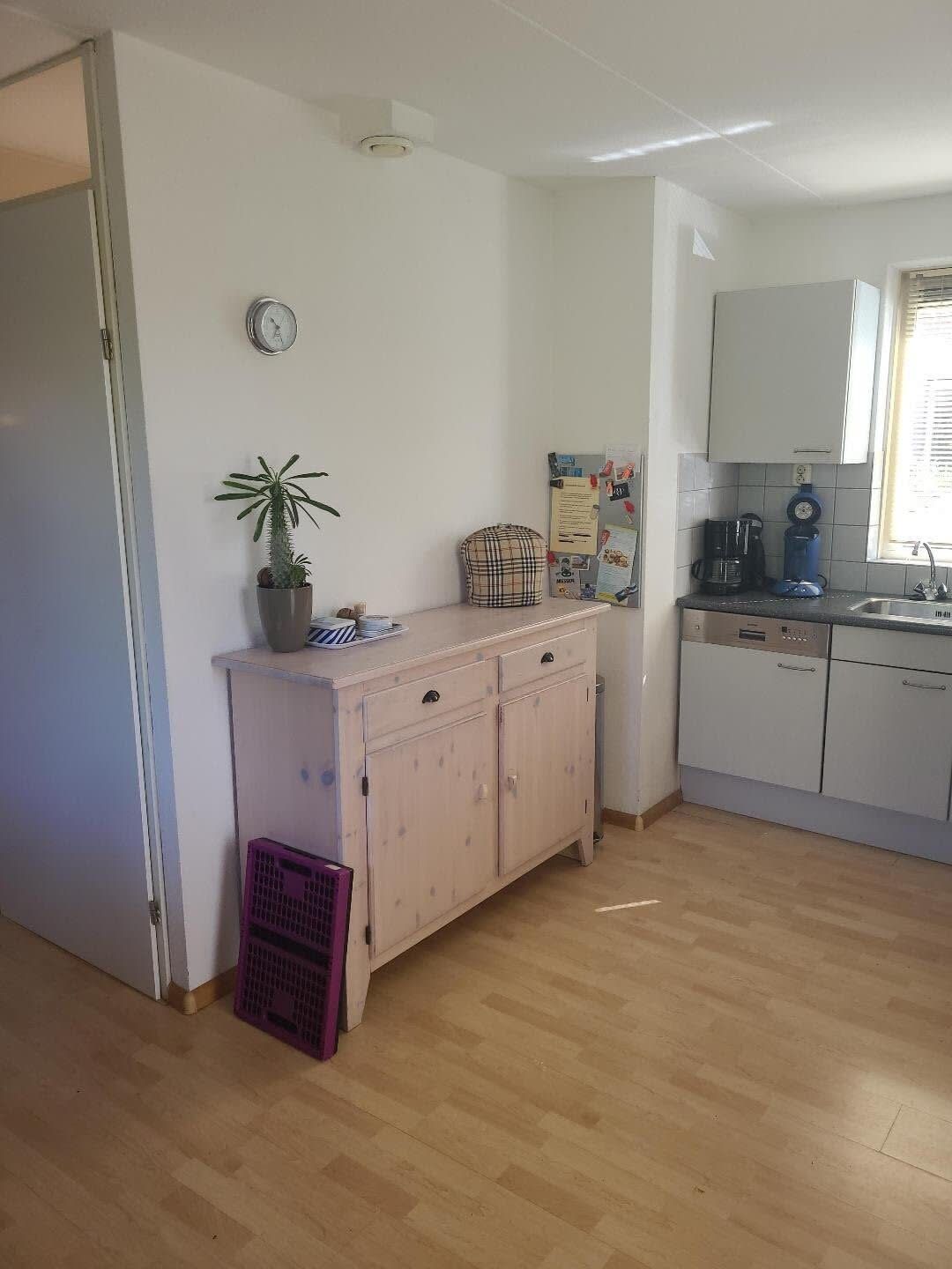 A compact kitchen with white cabinets, a small countertop, a clock, a potted plant, and a window with blinds.