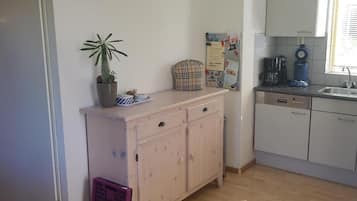 A compact kitchen with white cabinets, a small countertop, a clock, a potted plant, and a window with blinds.