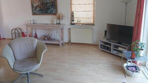 Ein Wohnzimmer mit Sofa, Fernseher, Heizkörper, Gemälde und einem Fenster mit Jalousien.