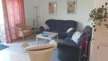 A living room with a dark blue sofa, a wicker chair, a coffee table, and a floor lamp. There are two framed pictures on the wall.