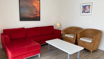 A red sofa, a white coffee table, and two wicker chairs in a room with a wooden floor and white walls.