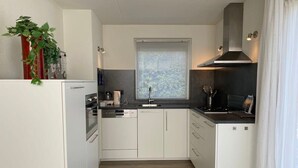 Une cuisine moderne avec des meubles blancs, un four encastré, une fenêtre avec des stores et une plante posée sur le plan de travail.