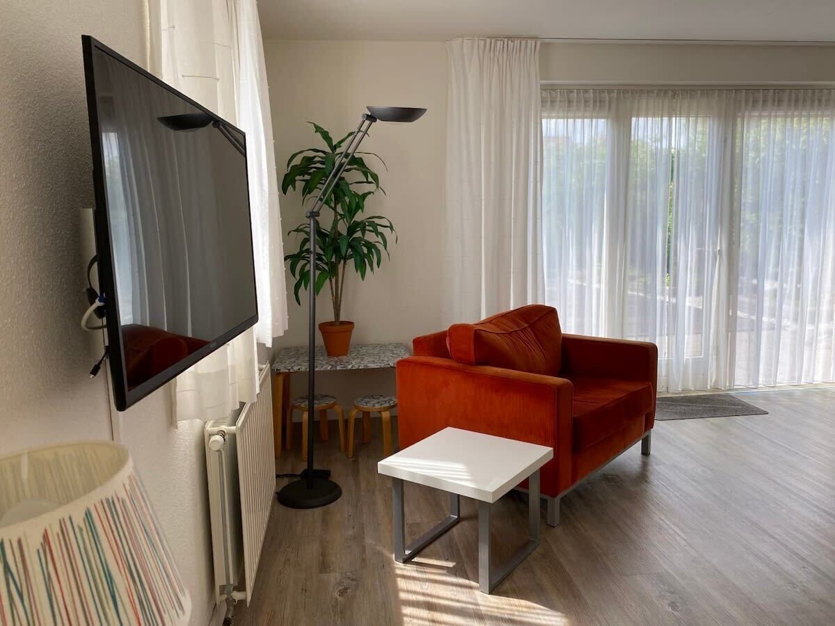 Un salon avec un téléviseur à écran plat, un canapé rouge, une petite table blanche, une plante en pot et une fenêtre ornée de rideaux légers.