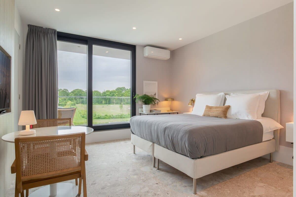 Un dormitorio moderno con una cama grande, un escritorio y una silla. Se ve vegetación por la ventana.