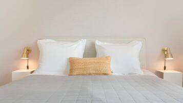 Una cama bien hecha con sábanas blancas y un almohadón texturizado, flanqueada por dos mesitas de noche con lámparas.