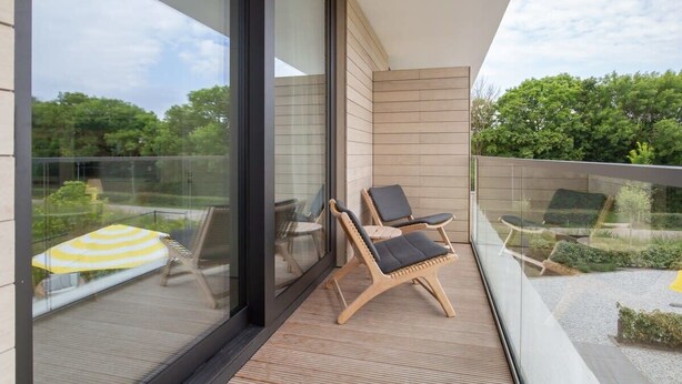 Un balcón con dos sillones de madera, barandilla de cristal y vista a zonas verdes.