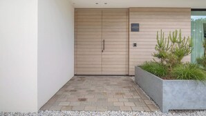 Entrada de un edificio moderno con puerta de madera, un letrero encima y una jardinera con plantas.