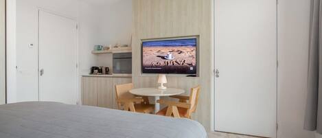 A bedroom with a bed, a small dining area with a table and chairs, a television mounted on a wall, and a kitchen area with cabinets and appliances.