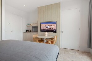 Ein Schlafzimmer mit einem Bett, einem kleinen Esstisch mit Stühlen, einem an der Wand befestigten Fernseher und einer Küchenzeile im Hintergrund.