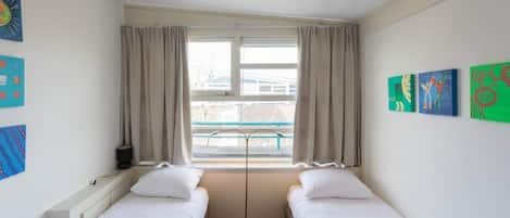 Una habitación con dos camas individuales, una ventana con cortinas y cuadros en las paredes.