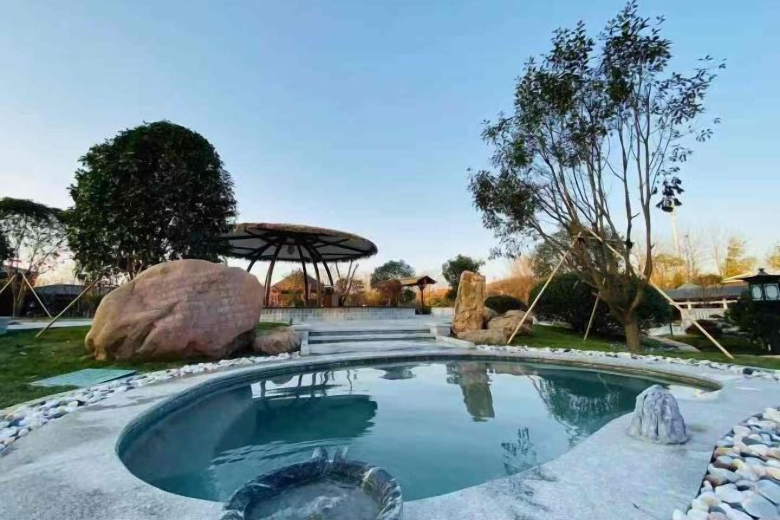 Une piscine avec un jacuzzi, entourée de rochers et d’arbres, et un espace salon couvert à l’arrière-plan.