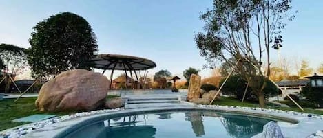A swimming pool with a hot tub, surrounded by rocks and trees, and a covered seating area in the background.