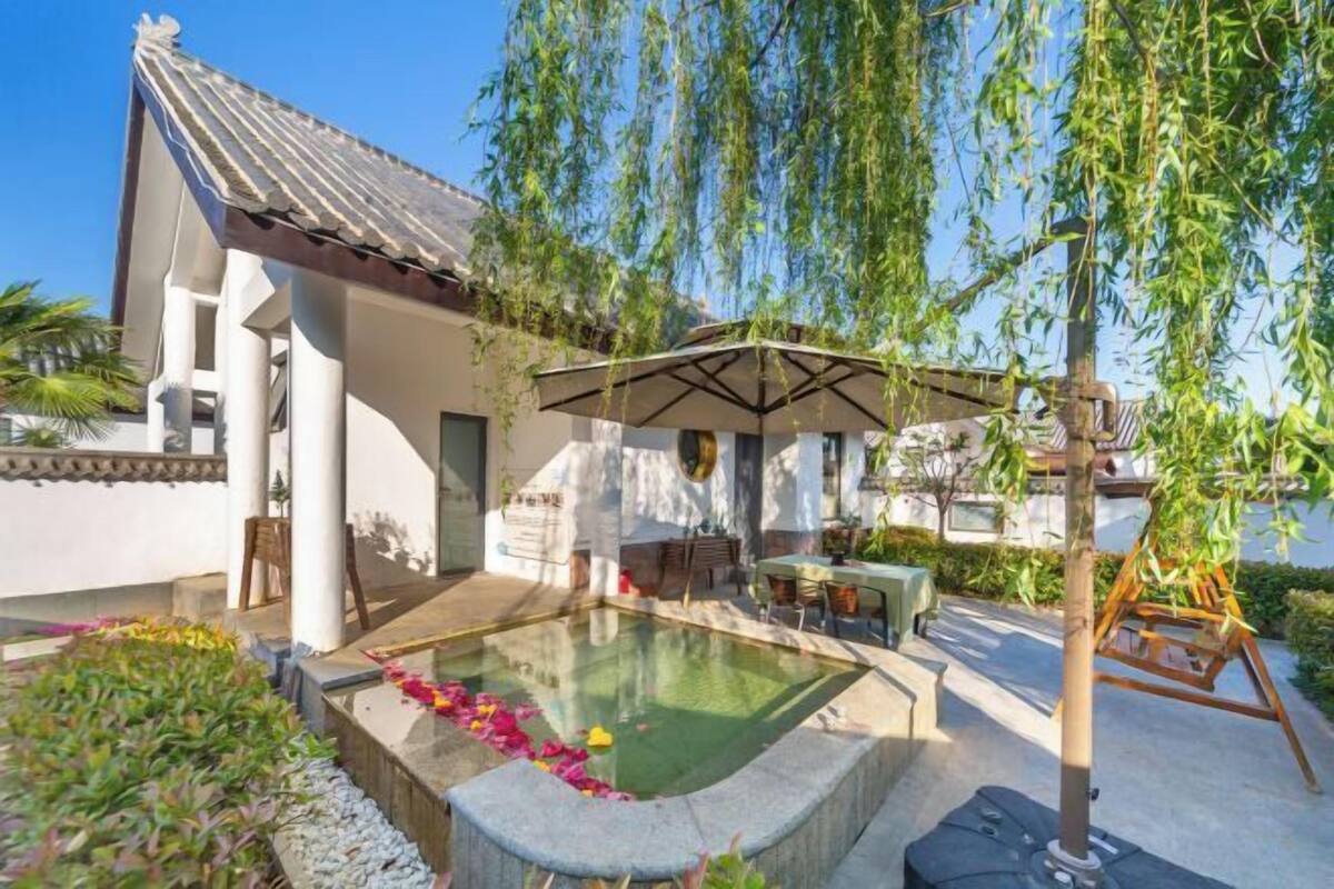 Un área de piscina con un pérgola, asientos y una pequeña construcción con techo de tejas.