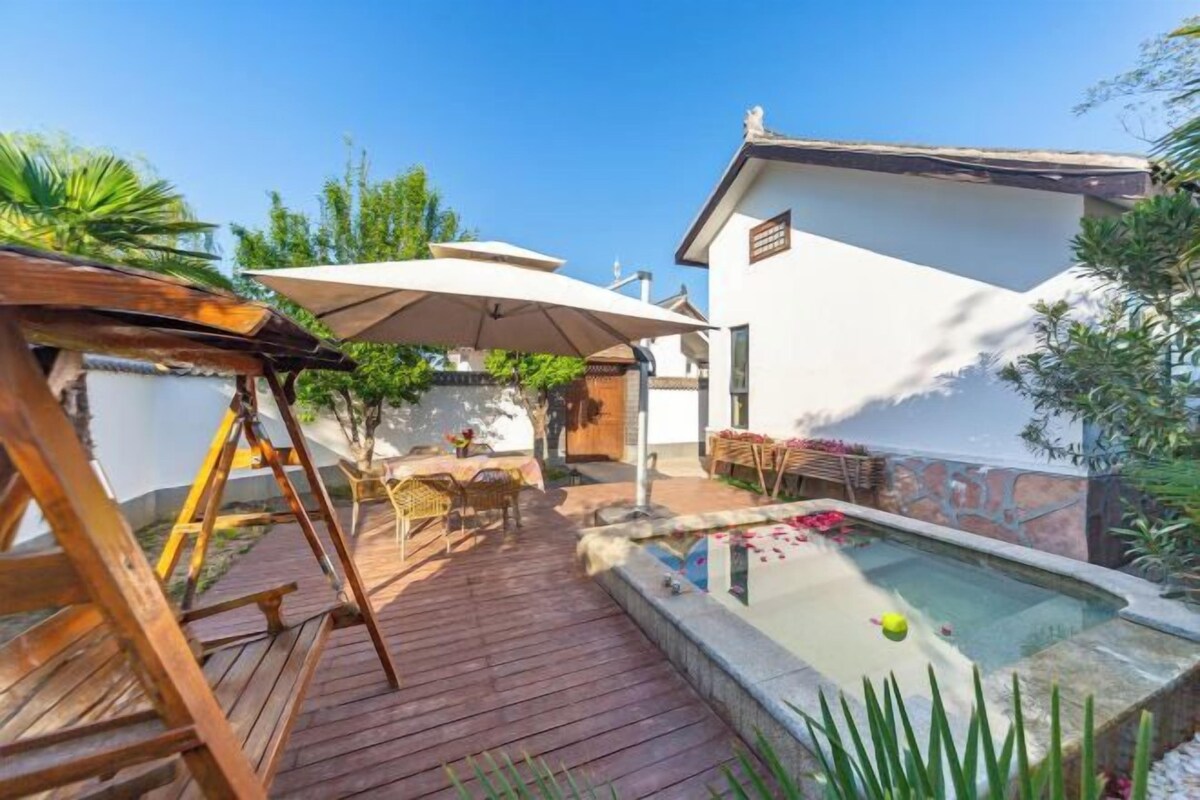 A wooden deck with outdoor furniture, a swimming pool, and a white building with a tiled roof.
