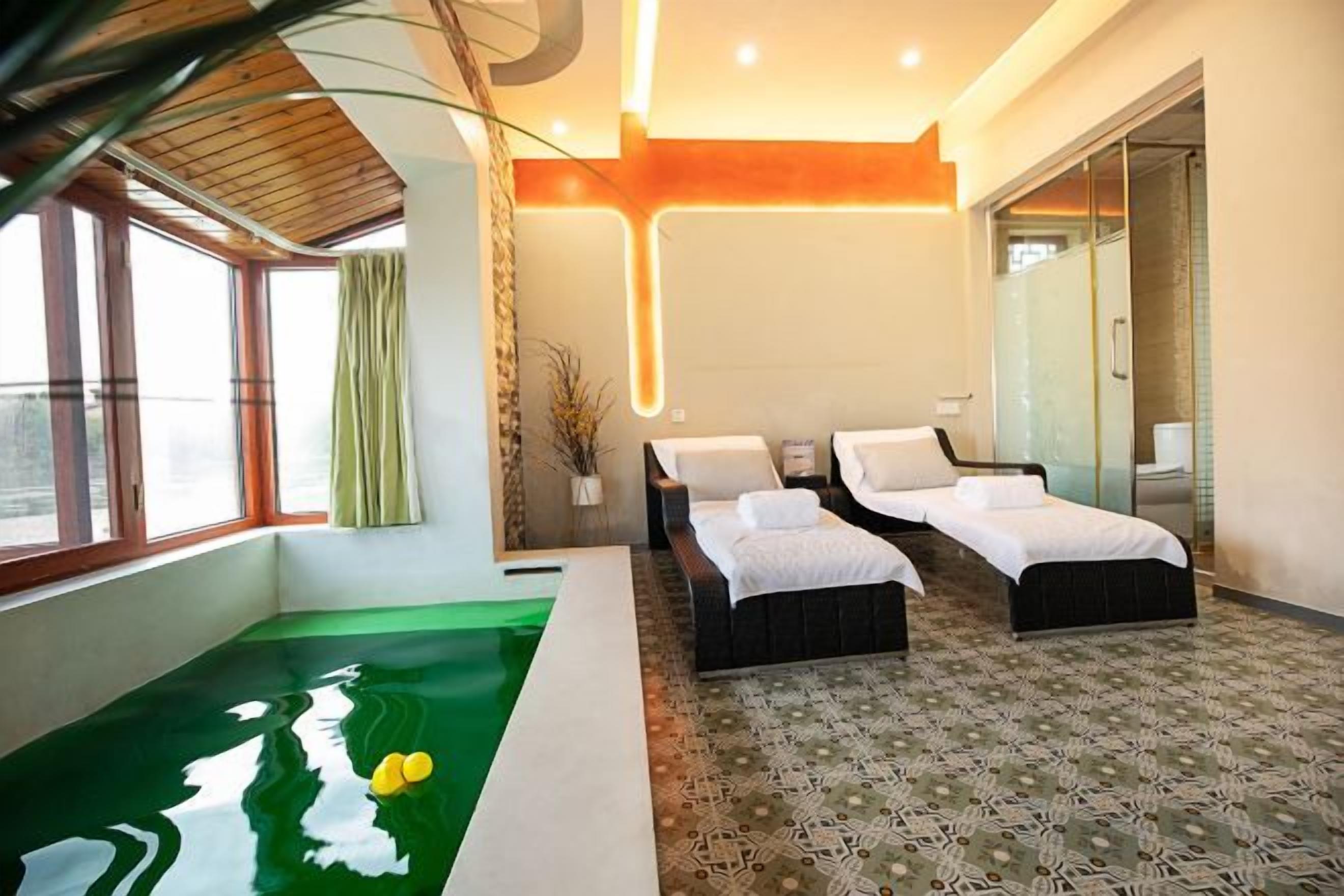 A hotel room with a large window, a green pool, two beds, and a glass door leading to a bathroom.