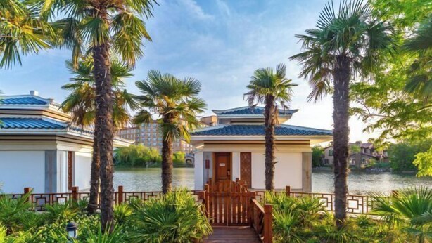 A hotel with a traditional architectural style, featuring a wooden gate and pathway leading to the entrance, surrounded by lush greenery and palm trees. - Yanling Bloom Hot Spring Hotel (Yanling)