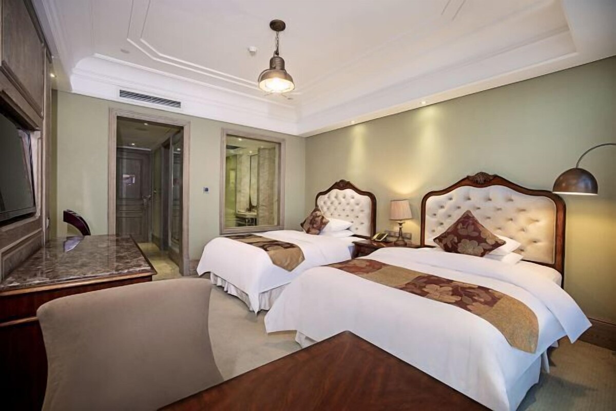 A hotel room with two beds, a desk, a chair, a lamp, and a ceiling light.