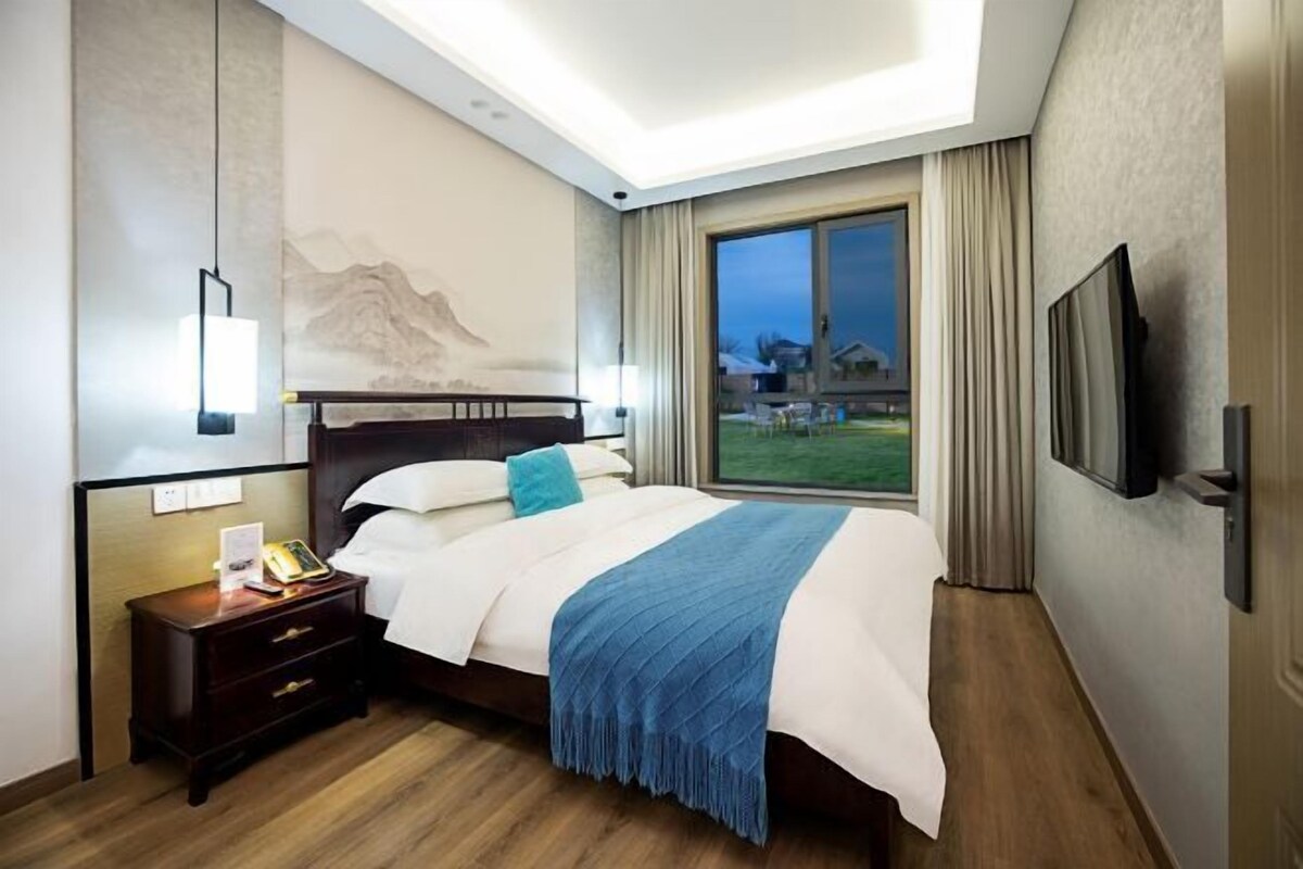 A modern hotel room with a large bed, a nightstand, a mounted TV, and a window with a view of a building and greenery.