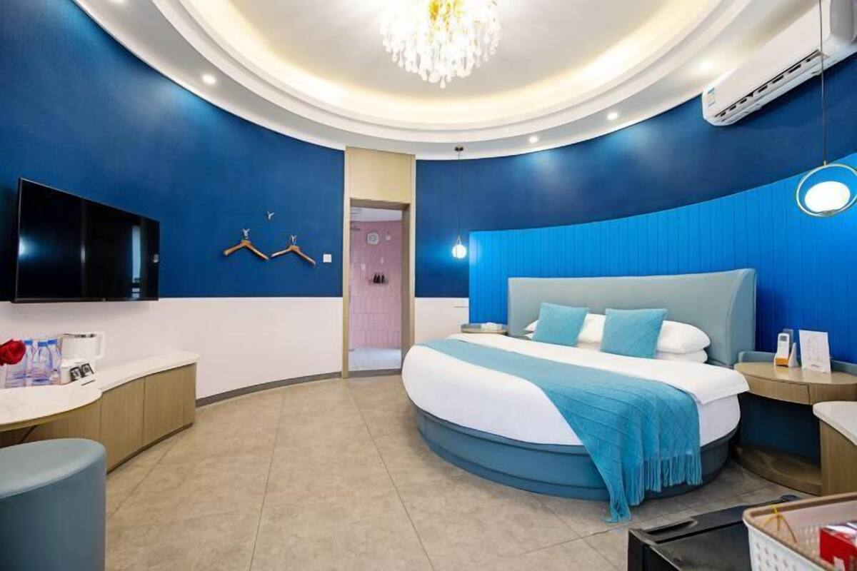 Una habitación de hotel moderna con techo curvo, una cama grande con ropa de cama azul, un televisor de pantalla plana, un escritorio con una silla y una pequeña mesa con una lámpara.