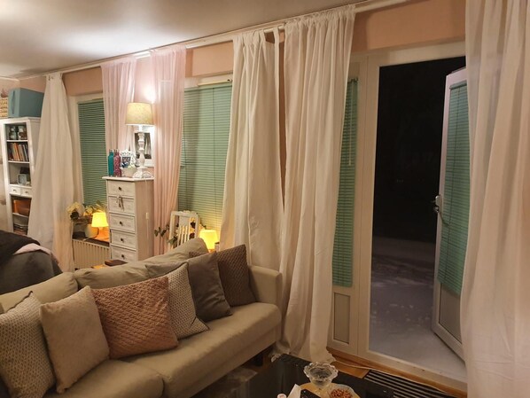 Un salón acogedor con un sofá, una mesita de noche con una lámpara y vistas al exterior a través de una ventana con contraventanas verdes.