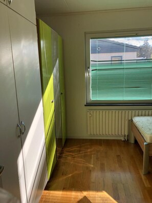 Una habitación con piso de madera, un radiador, una ventana con persianas y un armario con decoración verde y blanca.