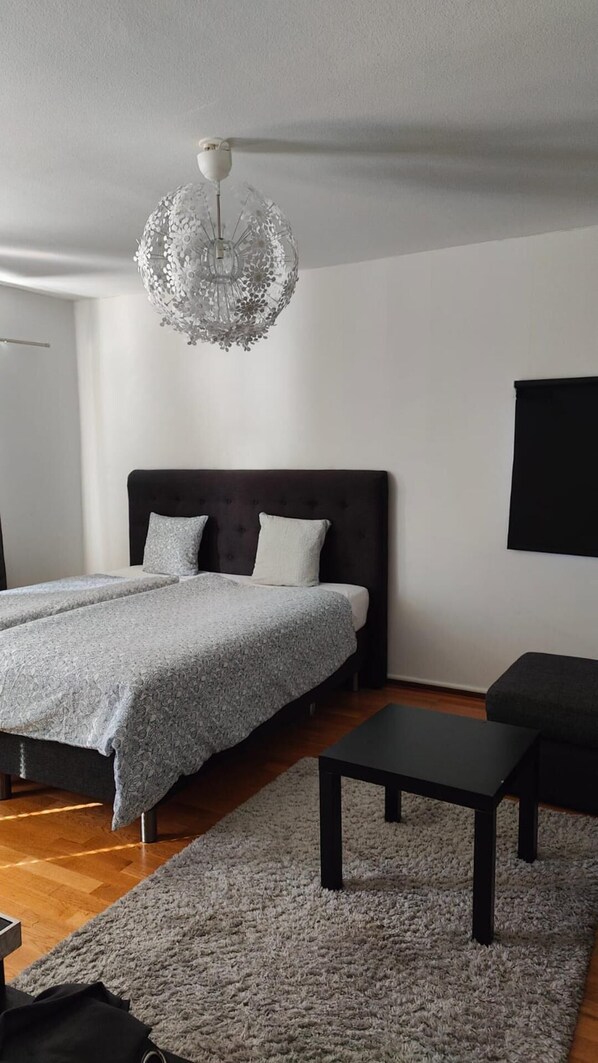 A modern bedroom with a large bed, a dark headboard, a grey patterned bedspread, a grey rug, a black coffee table, and a unique chandelier. - Very Nice Apartment 15 Minutes From Stockholm (Trangsund)