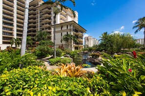 A hotel with multiple floors, a garden with various plants, a small pond, and a clear blue sky.