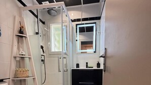 A compact bathroom with a glass-enclosed shower, a toilet, a sink with a mirror, and a small shelf with toiletries. - Charming studio 300 m from the beach of Santa Giulia (Porto-Vecchio)