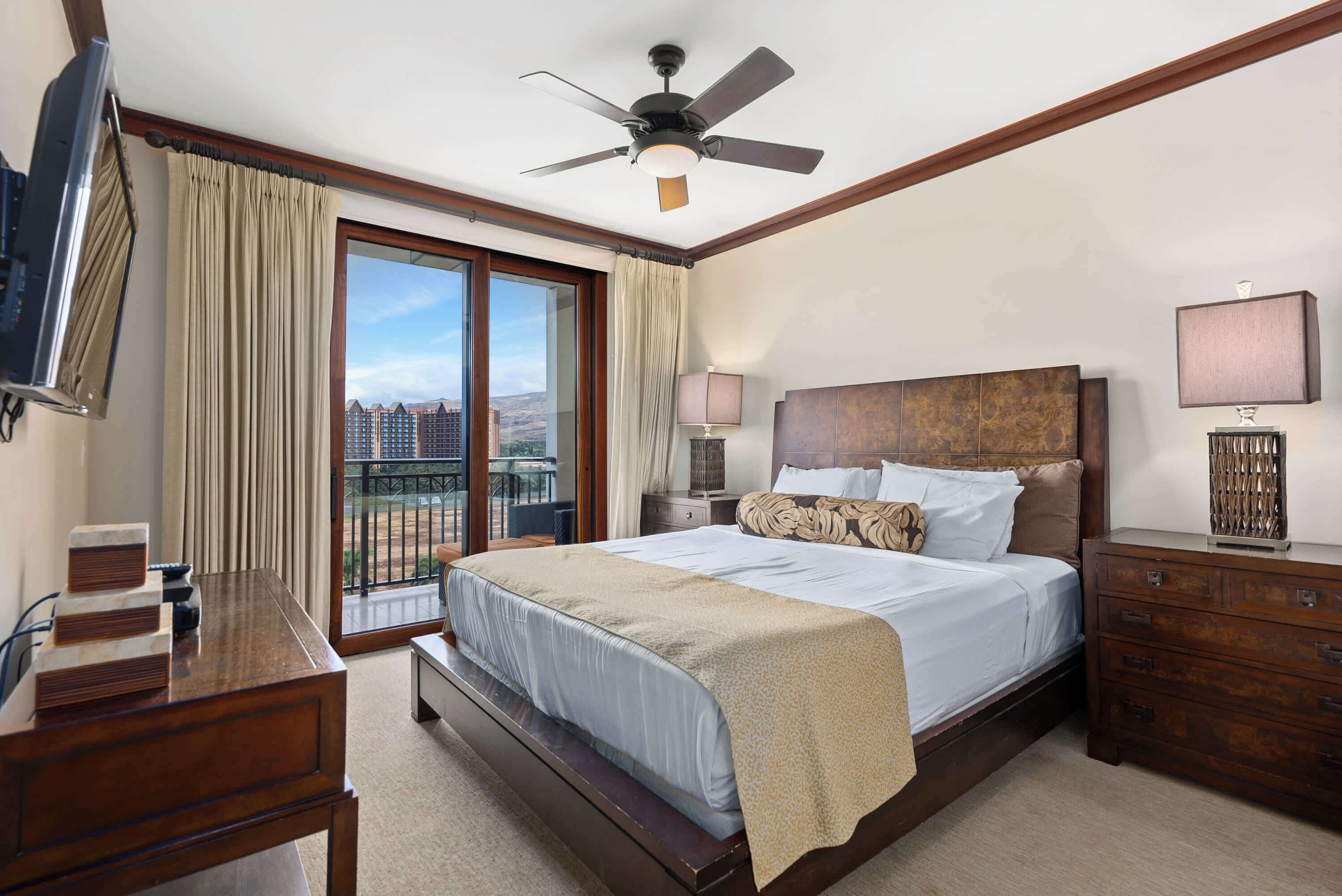 A hotel room with a bed, a desk, a TV, a ceiling fan, a nightstand, a lamp, and a view of the city.