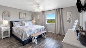 Un dormitorio con cama, mesitas de noche, ventilador de techo, ventana con cortinas y obras de arte en las paredes.