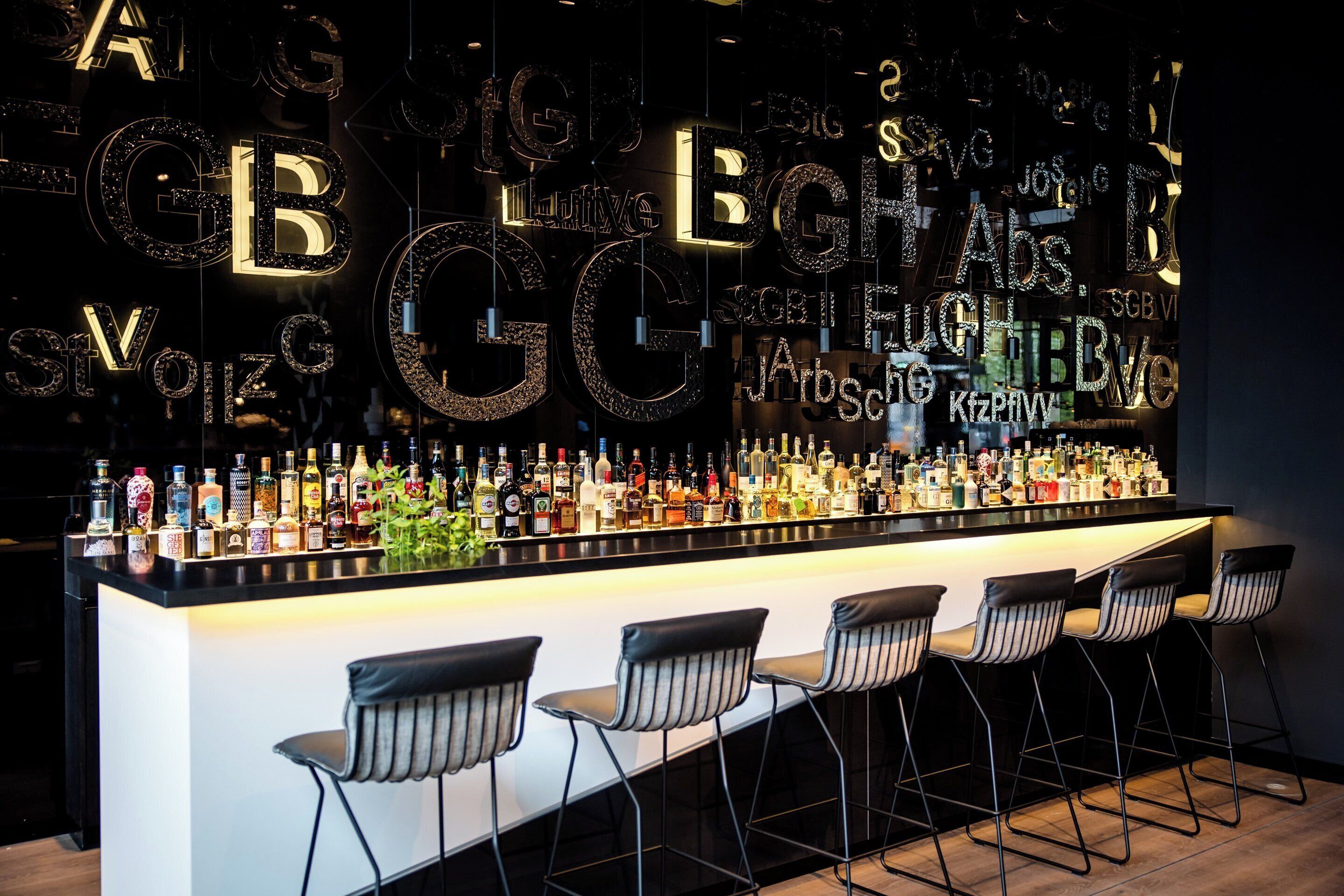 Una zona de bar con una barra alargada que exhibe varias botellas, y tres taburetes con rayas.
