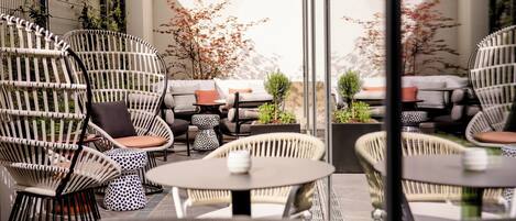 A modern outdoor seating area with wicker chairs and round tables, surrounded by a glass wall and plants.