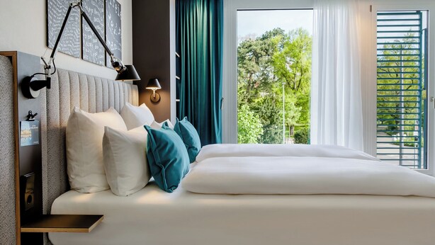 A modern hotel room with a large bed, a bedside table, a wall-mounted lamp, and a view of greenery outside. - Motel One Karlsruhe (Karlsruhe)