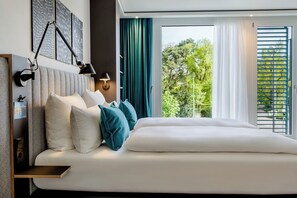 A modern hotel room with a large bed, a bedside table, a wall-mounted lamp, and a view of greenery outside. - Motel One Karlsruhe (Karlsruhe)