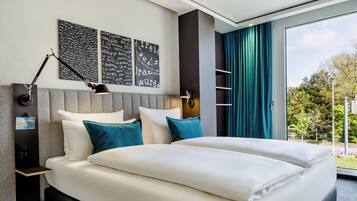 Un lit double avec des oreillers blancs et turquoise, une table de chevet avec une lampe et des œuvres d’art accrochées au mur.