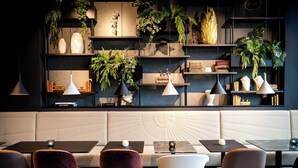 A modern interior with a seating area, tables, and shelves with plants and books. - Motel One Karlsruhe (Karlsruhe)