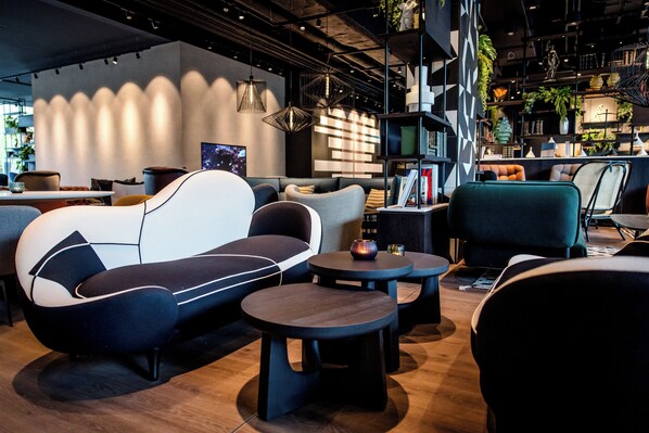 A modern lounge area with sofas, round tables, and a bookshelf. - Motel One Karlsruhe (Karlsruhe)