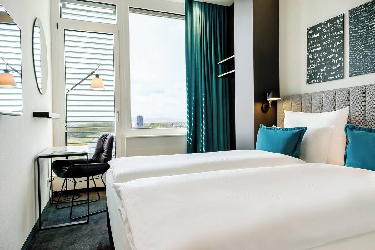 A hotel room with two beds, a desk with a chair, a window with blinds, and a view of the city.