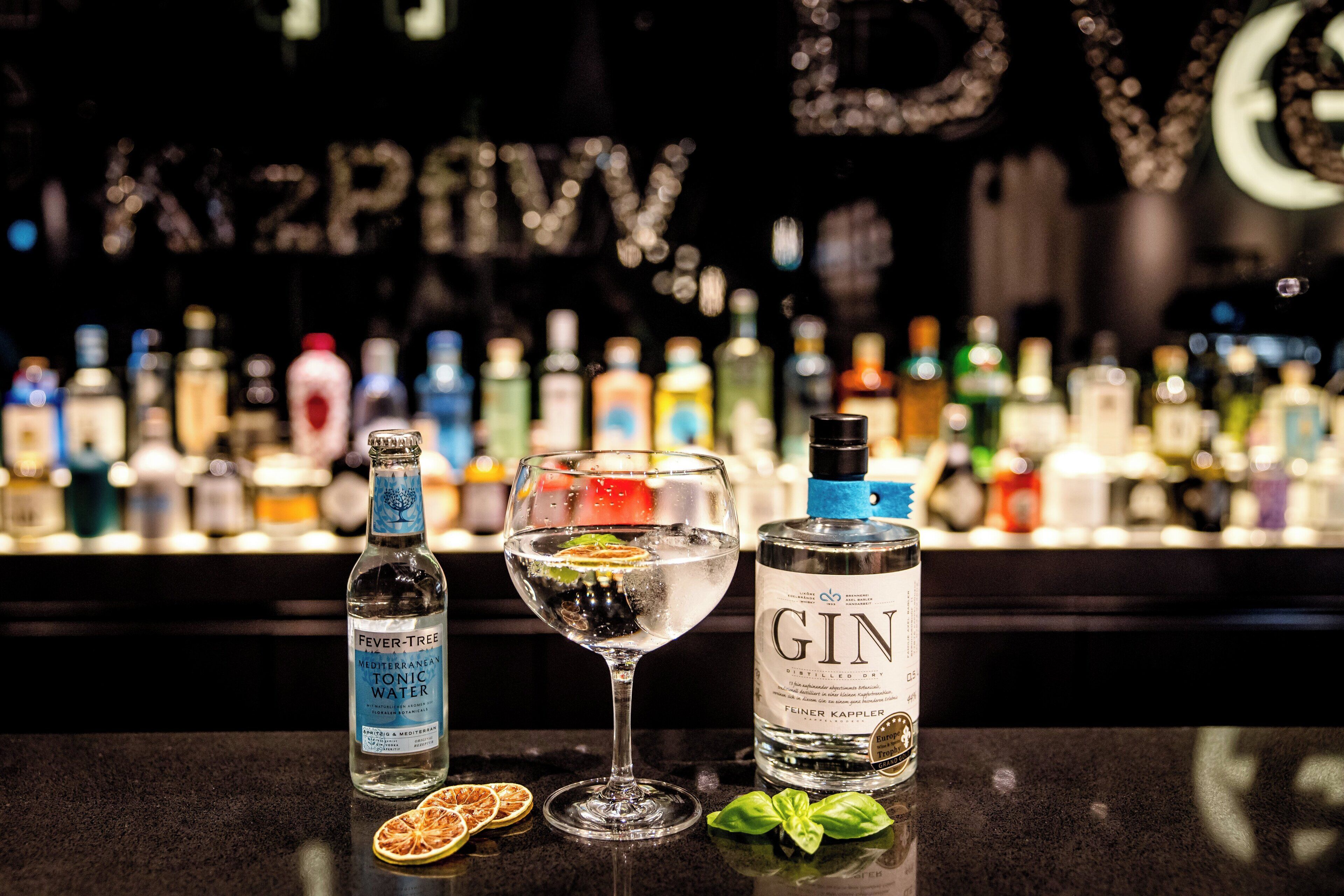 A bar setting with a glass of white wine, a bottle of gin, and a bottle of tonic water on a counter.