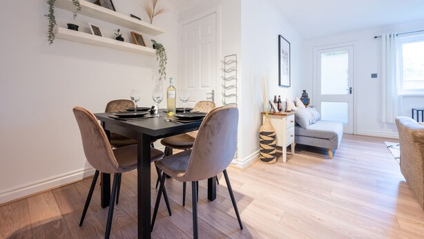 Een eetgedeelte met een zwarte tafel en vier stoelen, een witte deur, een grijze bank en een raam met gordijnen.