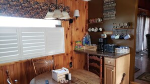 A wooden dining area with a round table, two chairs, a coffee maker, and a wall-mounted shelf with kitchenware.