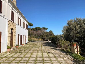 Une cour pavée, avec un bâtiment d’un côté, entourée de verdure et d’arbres.