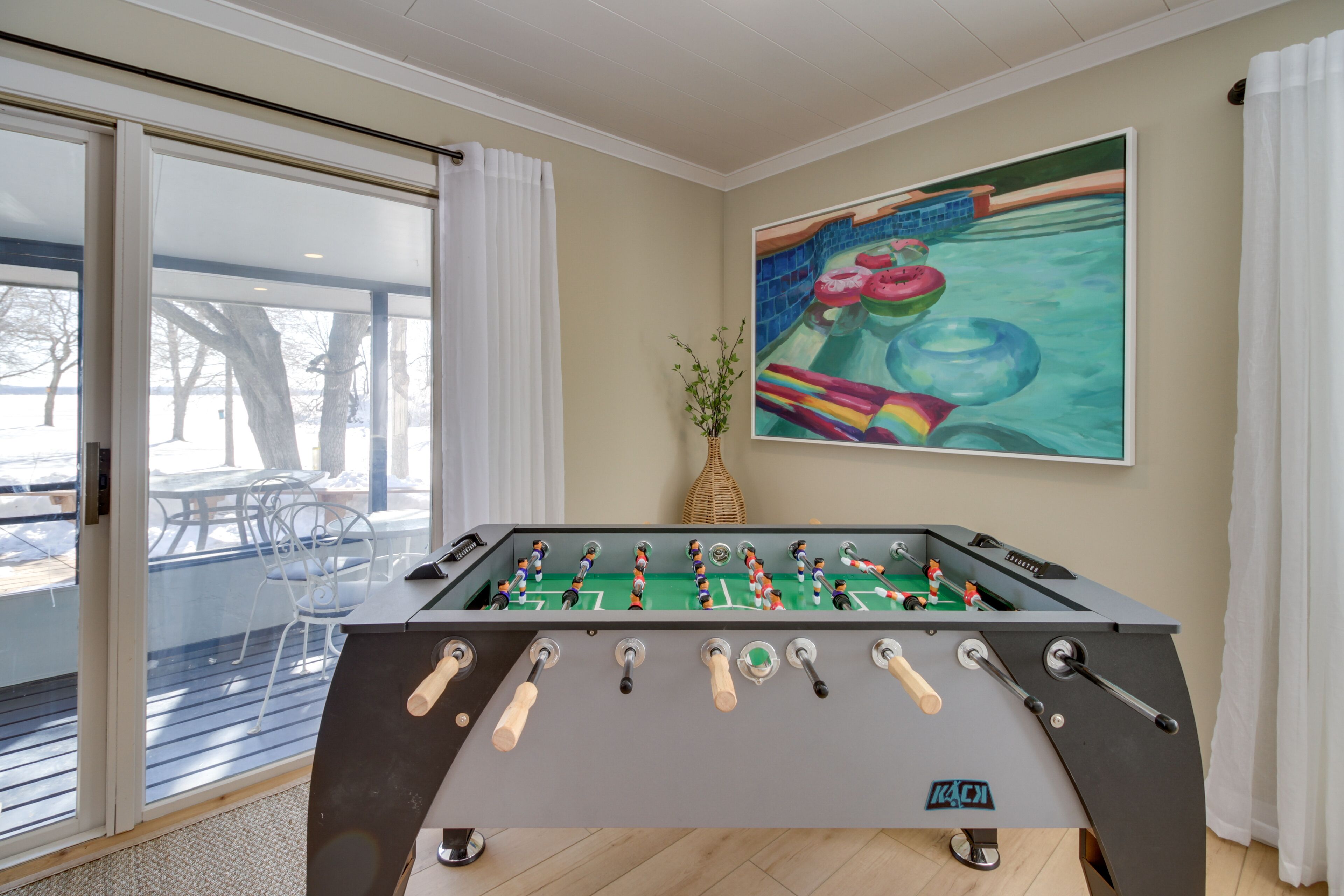 Een tafelvoetbaltafel in een kamer met een groot raam, een schilderij van een zwembad, en een rieten mand.
