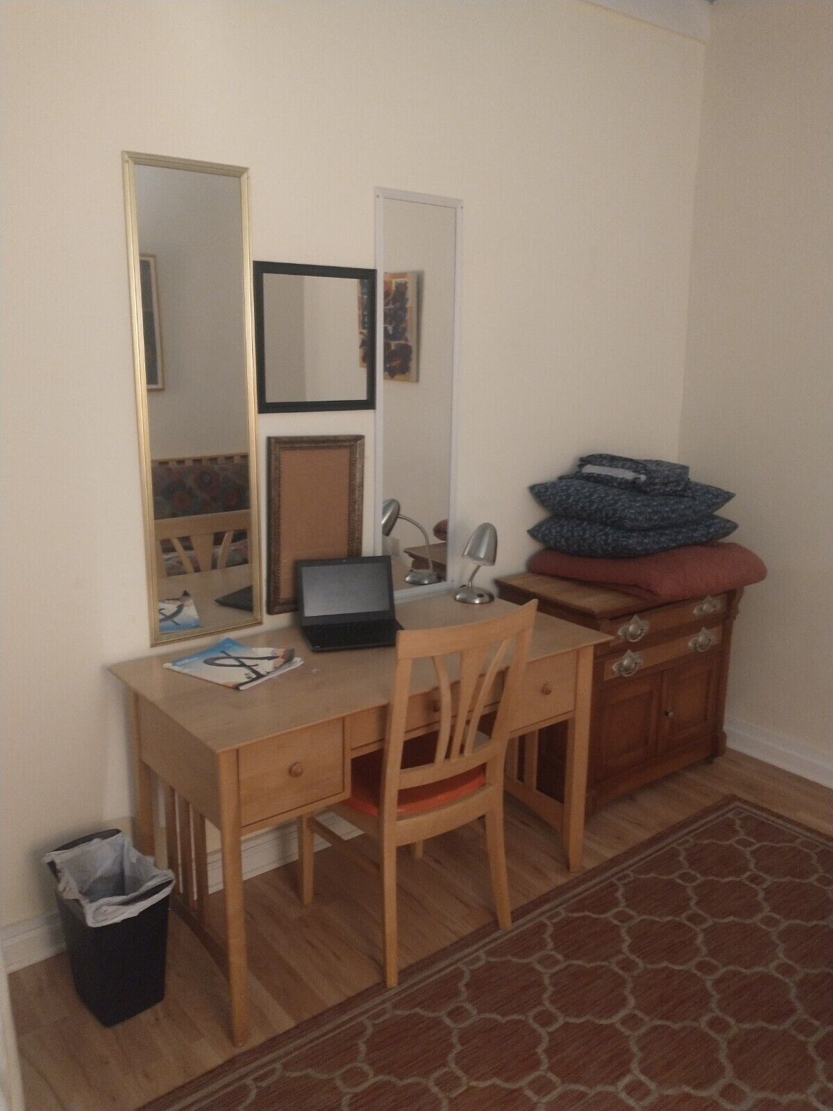 Una habitación pequeña y ordenada con un escritorio de madera, una silla, una computadora portátil, una lámpara y un espejo. Hay un cubo de basura pequeño y una alfombra estampada en el suelo.