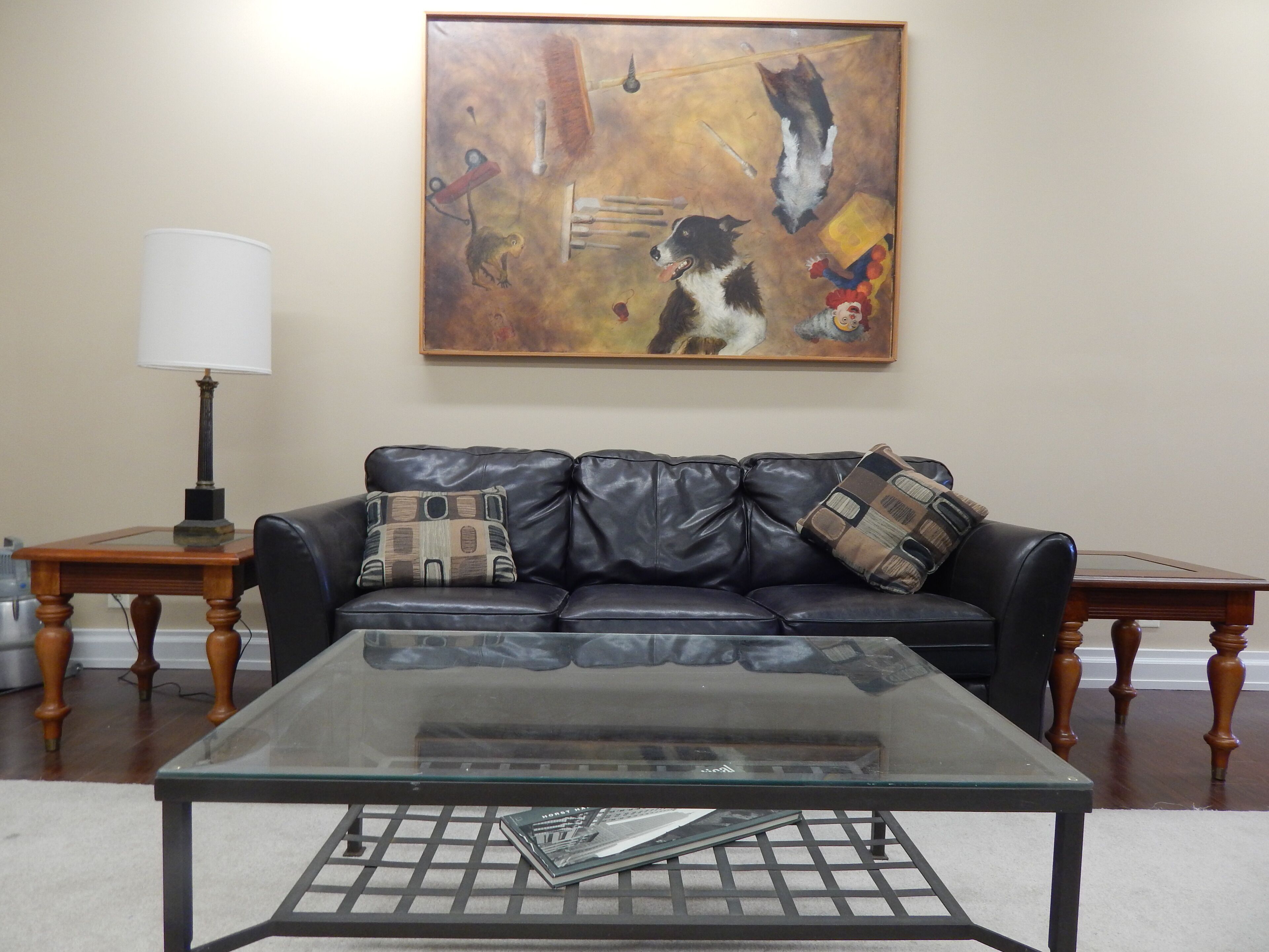 A living room with a glass coffee table, a black leather sofa, and a painting on the wall.