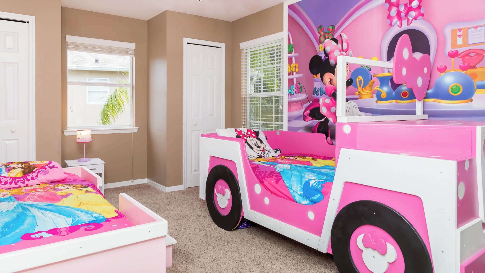 A child's bedroom with a pink and white playhouse, a bed with a Disney-themed bedspread, and a window with blinds.