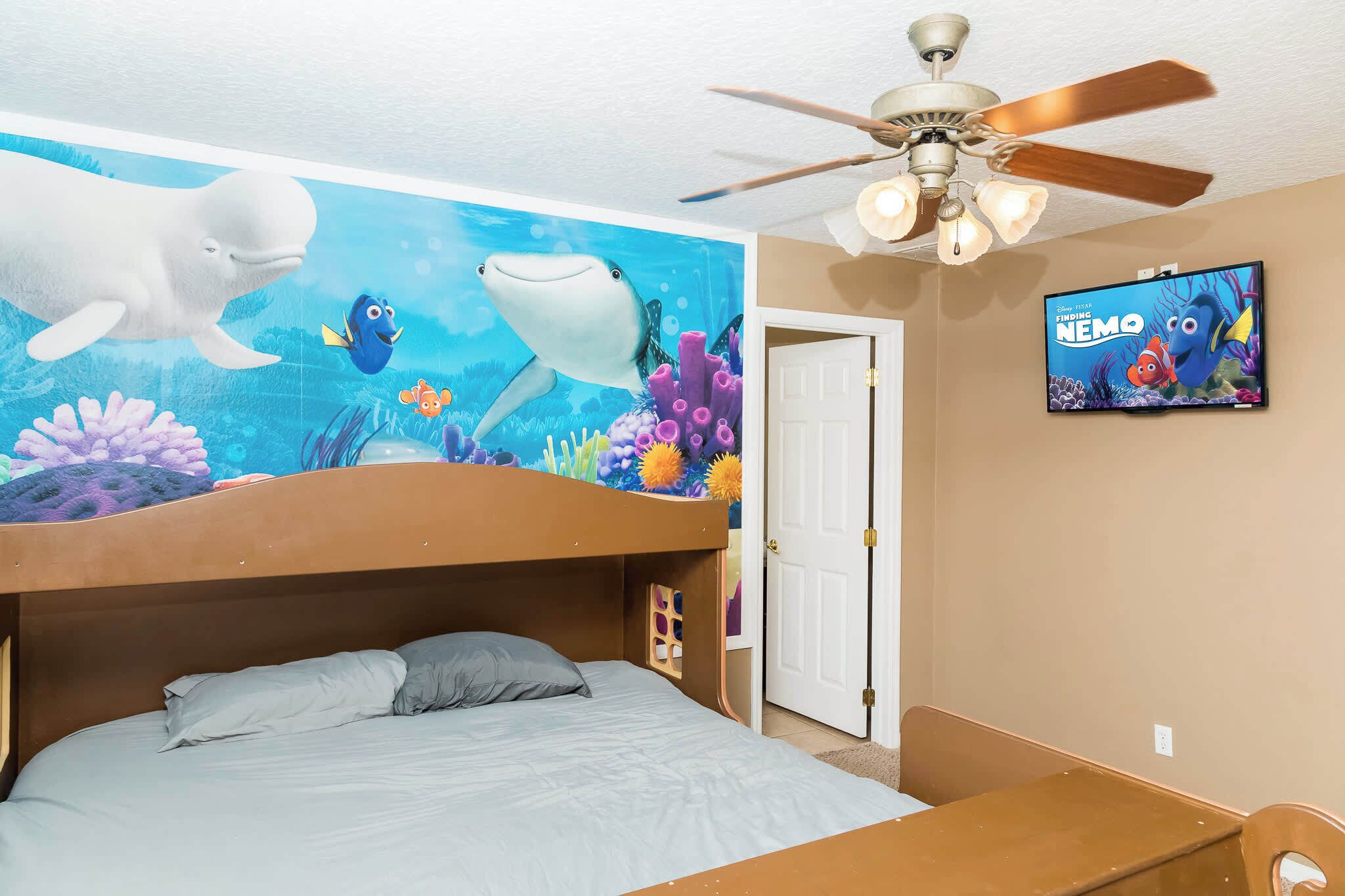 A bedroom with a bunk bed, a ceiling fan, a wall mural of an underwater scene, and a TV displaying a movie.